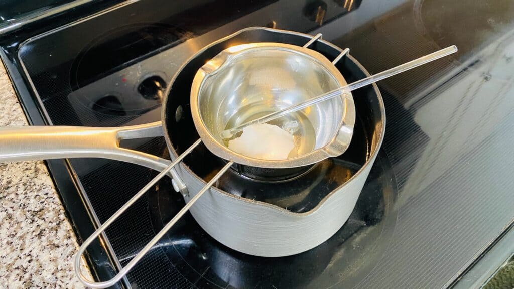 Adding coconut oil to the melted beeswax, glass stir rod resting inside the double boiler.